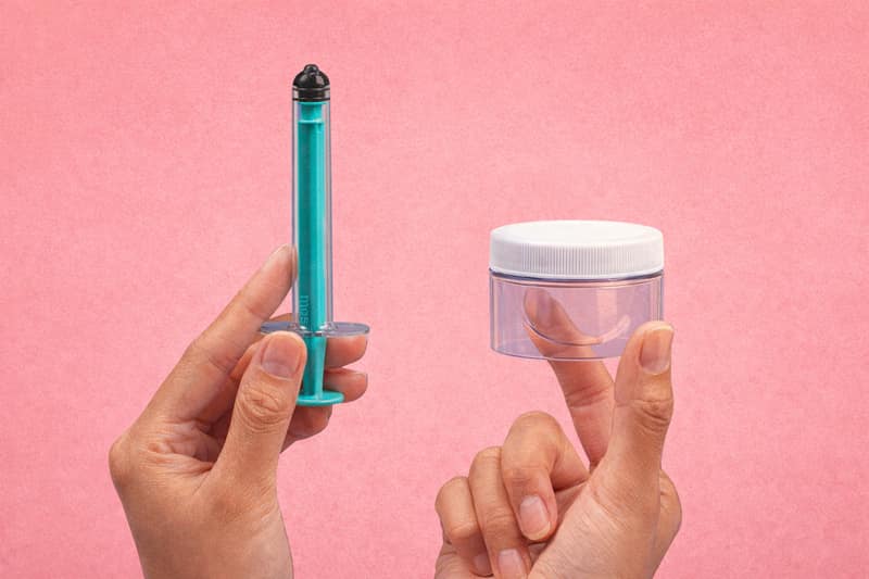 Hands holding syringe and collection cup on pink background showing home insemination kit tools for at-home conception