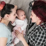 Two young women in casual clothes lying with their baby showing gay lesbian couples want a child through co-parenting or donor conception