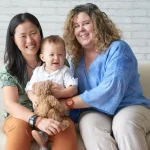 Lesbian IVF: same sex female couple with baby smiling together on sofa