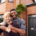 Portrait of excited gay male couple embracing outside their brick home celebrating same-sex adoption journey together