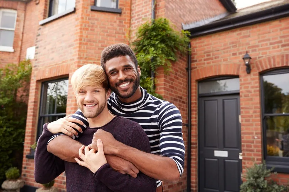 Portrait of excited gay male couple embracing outside their brick home celebrating same-sex adoption journey together