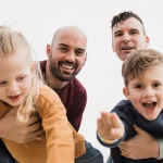 Gay male couple with little son and daughter playing together showing can gay people have kids through adoption or surrogacy