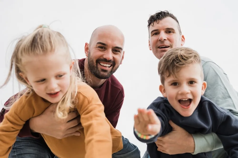 Gay male couple with little son and daughter playing together showing can gay people have kids through adoption or surrogacy